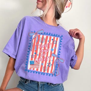 May include: A lilac-colored t-shirt with a patriotic design. The graphic features the text "Sweet Land of Liberty" within a frame with red and white stripes, stars, and an American flag. Blue bows and decorative swirls complete the design.