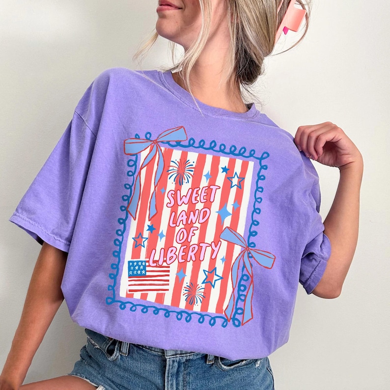 May include: A lilac-colored t-shirt with a patriotic design. The graphic features the text "Sweet Land of Liberty" within a frame with red and white stripes, stars, and an American flag. Blue bows and decorative swirls complete the design.