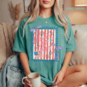May include: A light green t-shirt with a patriotic design. The design features red and white stripes, blue stars, and the text "Sweet Land of Liberty." The American flag and blue bows complete the design.
