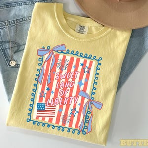 May include: A yellow t-shirt with a patriotic design featuring the text "Sweet Land of Liberty" in red and white, along with an American flag graphic and blue accents. The shirt is folded on top of blue jeans.