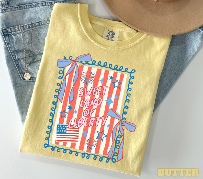May include: A yellow t-shirt with a patriotic design featuring the text "Sweet Land of Liberty" in red and white, along with an American flag graphic and blue accents. The shirt is folded on top of blue jeans.