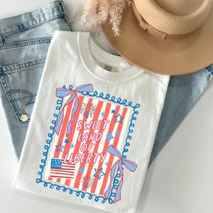 May include: White t-shirt with a patriotic design featuring red and white stripes, blue stars, and the text "Sweet Land of Liberty." The design includes a small American flag and blue bows. The shirt is paired with blue jeans and a tan hat.