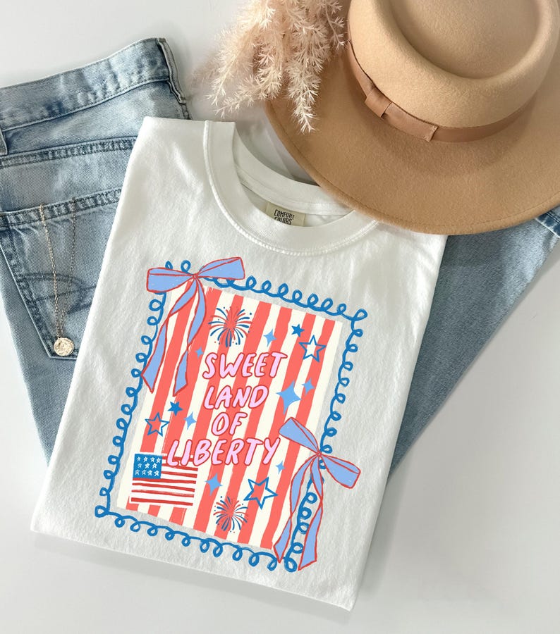 May include: White t-shirt with a patriotic design featuring red and white stripes, blue stars, and the text "Sweet Land of Liberty." The design includes a small American flag and blue bows. The shirt is paired with blue jeans and a tan hat.