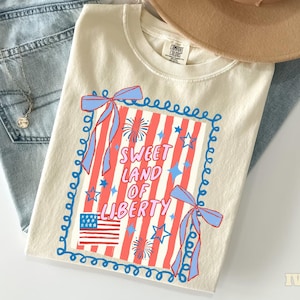 May include: Ivory-colored t-shirt with a patriotic design. The graphic features red and white stripes, a blue frame, bows, stars, fireworks, and the text "Sweet Land of Liberty." The shirt is paired with blue jeans and a tan hat.