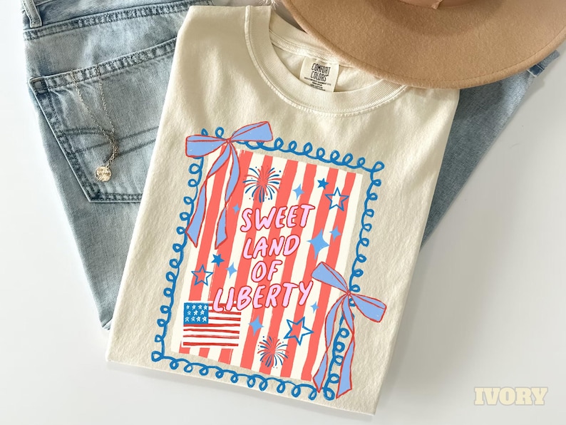 May include: Ivory-colored t-shirt with a patriotic design. The graphic features red and white stripes, a blue frame, bows, stars, fireworks, and the text "Sweet Land of Liberty." The shirt is paired with blue jeans and a tan hat.
