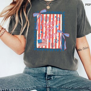 May include: Dark gray t-shirt featuring a patriotic design with the text "Sweet Land of Liberty." The graphic includes red and white stripes, blue stars, a small American flag, and decorative ribbons. The shirt is a relaxed fit.