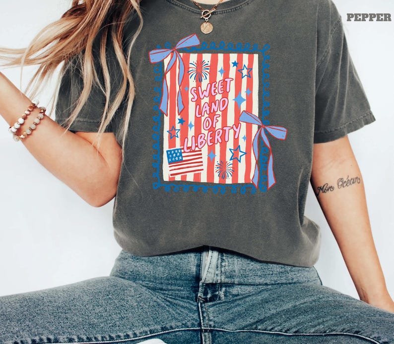 May include: Dark gray t-shirt featuring a patriotic design with the text "Sweet Land of Liberty." The graphic includes red and white stripes, blue stars, a small American flag, and decorative ribbons. The shirt is a relaxed fit.