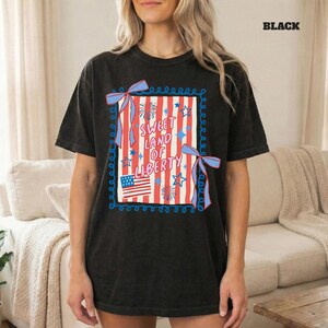 May include: Black t-shirt featuring a patriotic design. The graphic includes red and white stripes, blue stars, and the text "Sweet Land of Liberty." A small American flag is also present. The design is framed with a blue border and bows.