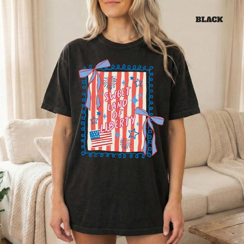 May include: Black t-shirt featuring a patriotic design. The graphic includes red and white stripes, blue stars, and the text "Sweet Land of Liberty." A small American flag is also present. The design is framed with a blue border and bows.