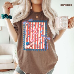 May include: Espresso-colored t-shirt featuring a patriotic design. The graphic includes the text "Sweet Land of Liberty" with an American flag, fireworks, and stars. The design is framed with blue ribbons and a decorative border.