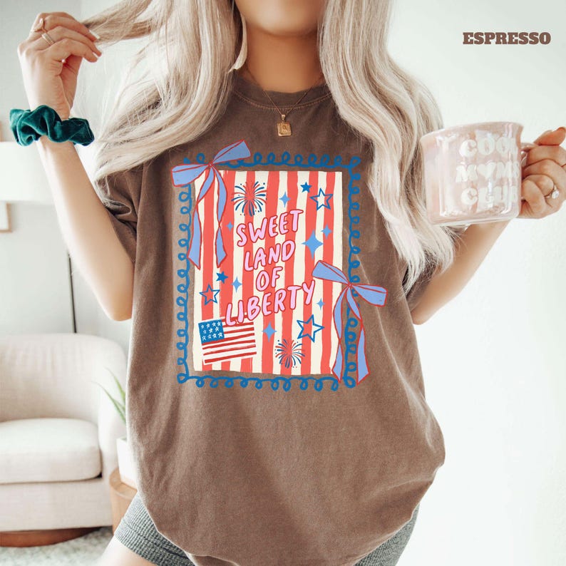 May include: Espresso-colored t-shirt featuring a patriotic design. The graphic includes the text "Sweet Land of Liberty" with an American flag, fireworks, and stars. The design is framed with blue ribbons and a decorative border.