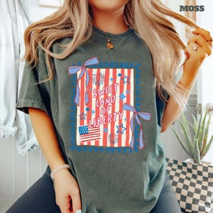 May include: A moss green t-shirt featuring a patriotic design. The design includes red and white stripes, blue stars, fireworks, and the text "Sweet Land of Liberty." Two blue bows frame the design, and an American flag is at the bottom.
