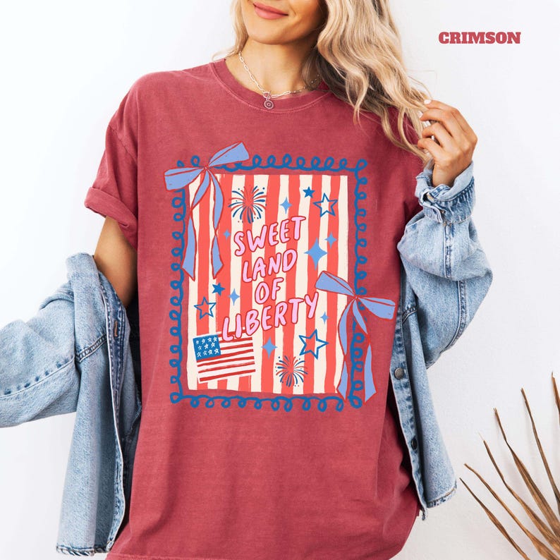 May include: Crimson-colored t-shirt with a patriotic design. The graphic features the text "Sweet Land of Liberty" in a playful font, along with an American flag, stars, and fireworks. The design is framed with a blue border and ribbons.