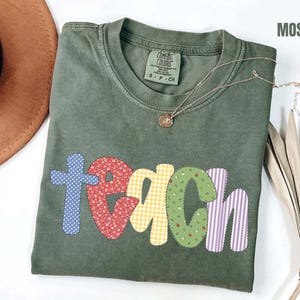 Pode incluir: Uma t-shirt de manga curta verde-musgo com decote redondo. A palavra "teach" está impressa na frente em letras grandes, coloridas e estampadas. Cada letra apresenta um design diferente, incluindo bolinhas azuis, flores vermelhas, xadrez vichy amarelo, estampa de maçãs verdes e listras roxas.