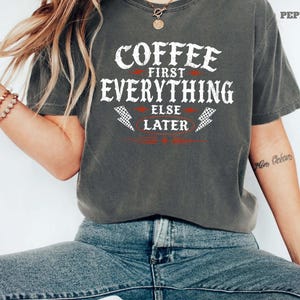 May include: Dark gray t-shirt with the text "COFFEE FIRST EVERYTHING ELSE LATER" in white, with red accents and lightning bolt graphics. The shirt is worn by a person wearing blue jeans.