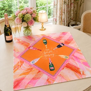 May include: A round table setting with a pink and orange patterned table runner. The runner features champagne bottles and the text "Sip, Sparkle, Mahjong." A bottle of Mo&euml;t champagne, two glasses, and a bouquet of pink roses are also on the table.