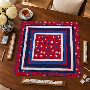 May include: A square game mat featuring a red background with scattered white and blue stars, framed by concentric borders in white, blue, and red. The mat is positioned on a wooden table with mahjong tiles and racks arranged around it.