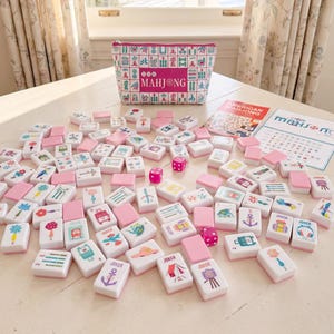 May include: A Mahjong game set featuring white tiles with colorful illustrations and pink backs, a matching zippered storage pouch with a Mahjong pattern, three pink dice, and two instructional cards with the text American Mahjong and Mahjong.