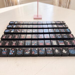 May include: A collection of black Mahjong tiles, each displaying unique symbols and characters in pink, blue, and white. The tiles are neatly arranged, highlighting the game's components. The image captures the essence of the classic tile-based game.