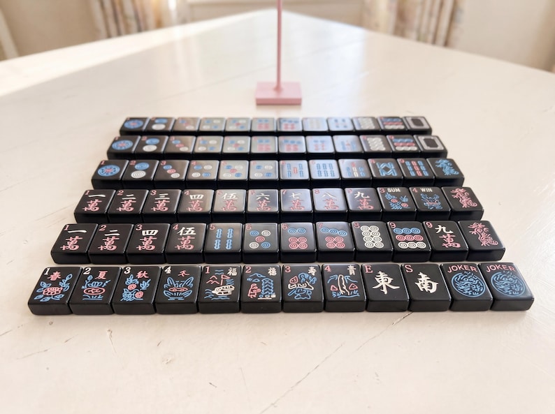 May include: A collection of black Mahjong tiles, each displaying unique symbols and characters in pink, blue, and white. The tiles are neatly arranged, highlighting the game's components. The image captures the essence of the classic tile-based game.