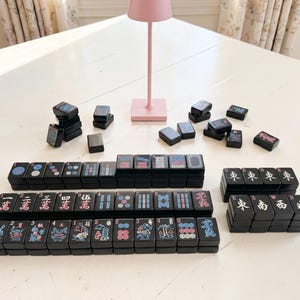 May include: A black Mahjong set with colourful symbols and characters. The tiles are arranged in stacks and rows, with some scattered around. A pink lamp is in the background.