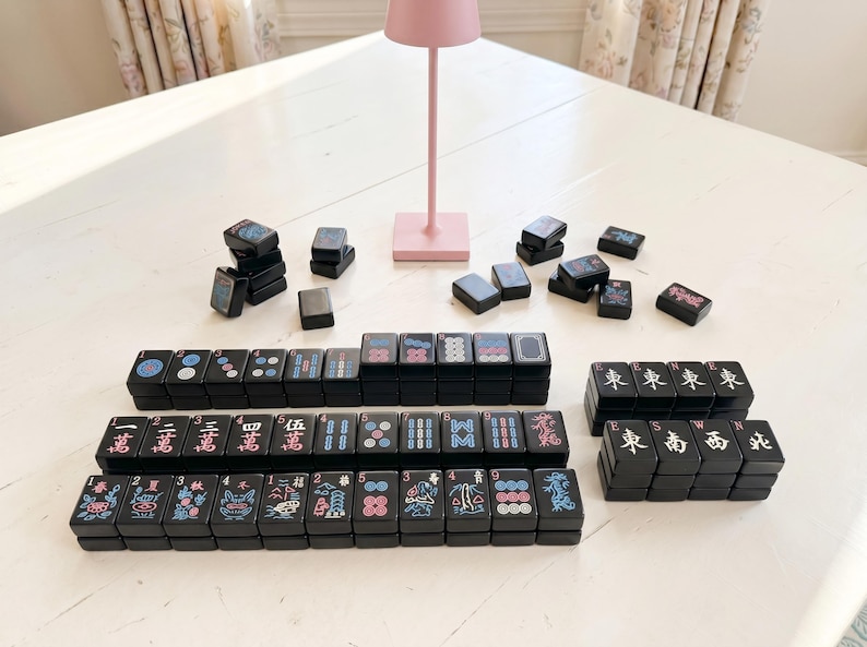 May include: A black Mahjong set with colourful symbols and characters. The tiles are arranged in stacks and rows, with some scattered around. A pink lamp is in the background.