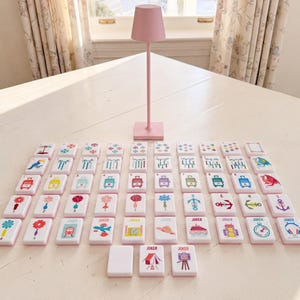May include: A set of rectangular white tiles featuring colorful graphic illustrations of various objects, symbols, and animals, arranged in a grid pattern on a surface, accompanied by a tall, slender, solid light pink table lamp with a conical shade.