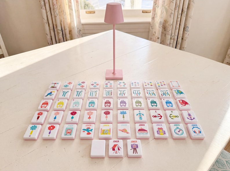 May include: A set of rectangular white tiles featuring colorful graphic illustrations of various objects, symbols, and animals, arranged in a grid pattern on a surface, accompanied by a tall, slender, solid light pink table lamp with a conical shade.