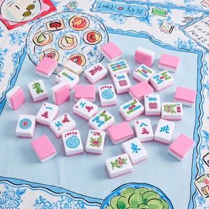 May include: A collection of Mahjong tiles with white faces and pink backs scattered on a light blue fabric surface. The tiles feature various colorful symbols, Chinese characters, and floral designs, including specific tiles labeled Joker and WIN.