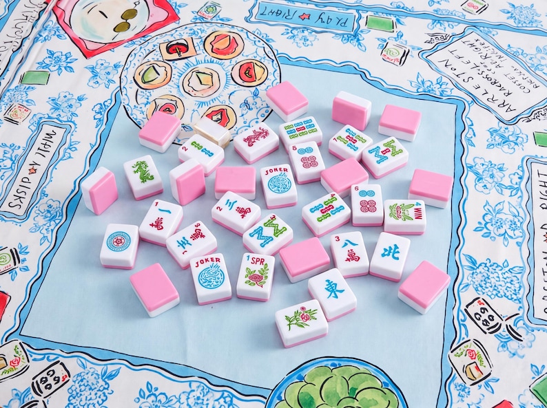 May include: A collection of Mahjong tiles with white faces and pink backs scattered on a light blue fabric surface. The tiles feature various colorful symbols, Chinese characters, and floral designs, including specific tiles labeled Joker and WIN.
