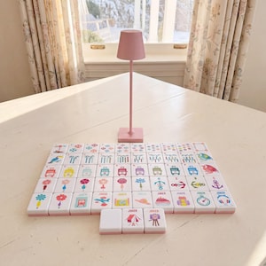 May include: A set of white mahjong tiles with colorful graphic illustrations including flowers, animals, and travel icons, arranged in a grid pattern. A tall, slender, light pink lamp with a conical shade stands behind the tiles.