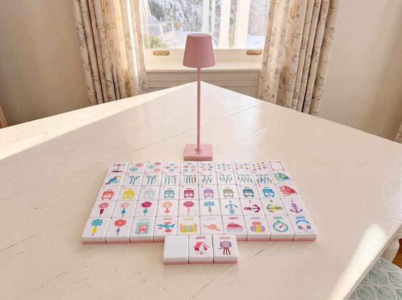 May include: A set of white mahjong tiles with colorful graphic illustrations including flowers, animals, and travel icons, arranged in a grid pattern. A tall, slender, light pink lamp with a conical shade stands behind the tiles.