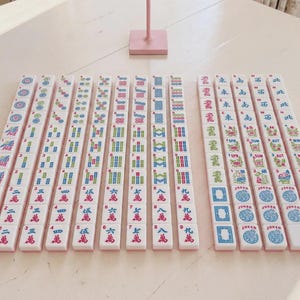 May include: A set of white mahjong tiles arranged in rows on a light surface. The tiles feature colorful engraved symbols, Chinese characters, numbers, and English text including Joker, Spr, Sum, Aut, and Win. One pink tile is at the end of a row.