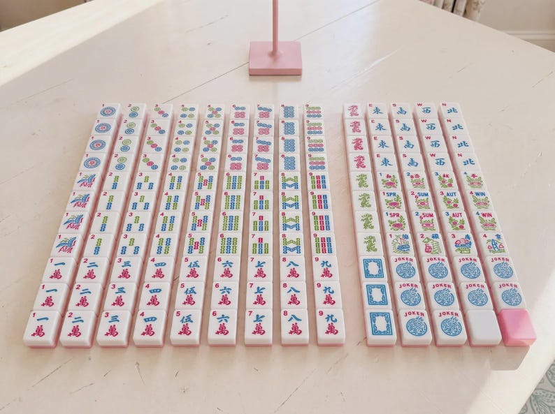 May include: A set of white mahjong tiles arranged in rows on a light surface. The tiles feature colorful engraved symbols, Chinese characters, numbers, and English text including Joker, Spr, Sum, Aut, and Win. One pink tile is at the end of a row.