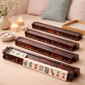 以下が含まれることがあります： 茶色と琥珀色の麻雀牌ラック4つと、それに合う牌のセット。牌は白地に赤、緑、黒のシンボル。ラックはべっ甲模様です。麻雀セットです。