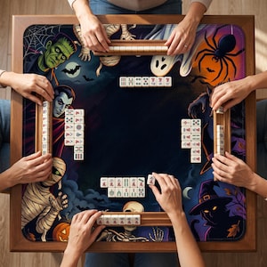 May include: A square mahjong table featuring a Halloween-themed mat with illustrations of Frankenstein, a vampire, a mummy, a skeleton, a witch, a ghost, and a spider. Four wooden tile racks hold white mahjong tiles, with additional tiles arranged on the mat.