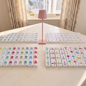 May include: A set of white rectangular mahjong tiles arranged in four distinct groups on a table, featuring colorful printed icons such as suitcases, anchors, flowers, and dragons, accompanied by a small, solid pink metal table lamp in the center.
