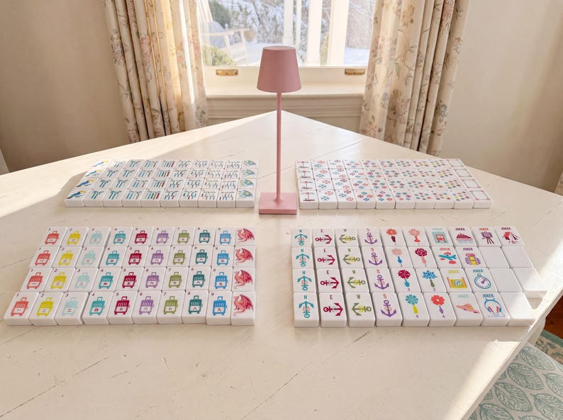 May include: A set of white rectangular mahjong tiles arranged in four distinct groups on a table, featuring colorful printed icons such as suitcases, anchors, flowers, and dragons, accompanied by a small, solid pink metal table lamp in the center.