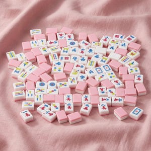 May include: A collection of pink and white Mahjong tiles scattered on a pink fabric surface. The tiles feature various symbols and characters in red, blue, and green. The pink tiles are rectangular and serve as the base for the white tiles.
