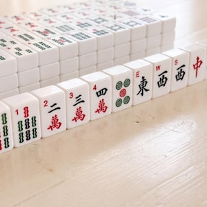 May include: A collection of white Mahjong tiles with red, green, and black markings. The tiles are arranged in stacks and rows, with some tiles displaying numbers, Chinese characters, and symbols. The game pieces are on a light-colored wooden surface.