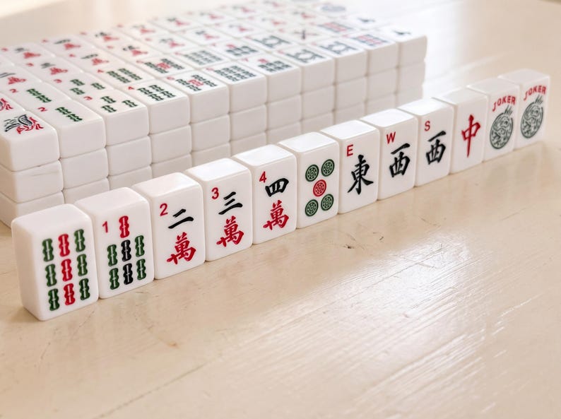 May include: A collection of white Mahjong tiles with red, green, and black markings. The tiles are arranged in stacks and rows, with some tiles displaying numbers, Chinese characters, and symbols. The game pieces are on a light-colored wooden surface.