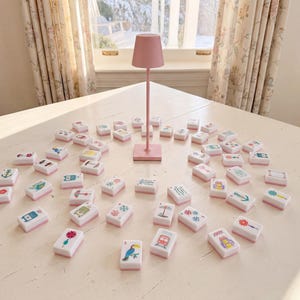 May include: A set of white rectangular mahjong tiles with colorful graphic illustrations on the faces, arranged in a circular pattern around a tall, slender, light pink metal lamp with a conical shade standing on a square base.