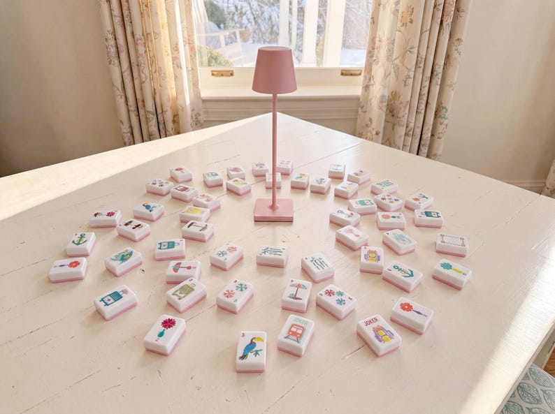 May include: A set of white rectangular mahjong tiles with colorful graphic illustrations on the faces, arranged in a circular pattern around a tall, slender, light pink metal lamp with a conical shade standing on a square base.
