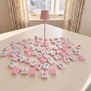 May include: A set of rectangular mahjong tiles with white faces featuring various colorful illustrations and pink backs, arranged in a scattered pattern around a tall, slender, solid pink table lamp with a conical shade on a white surface.