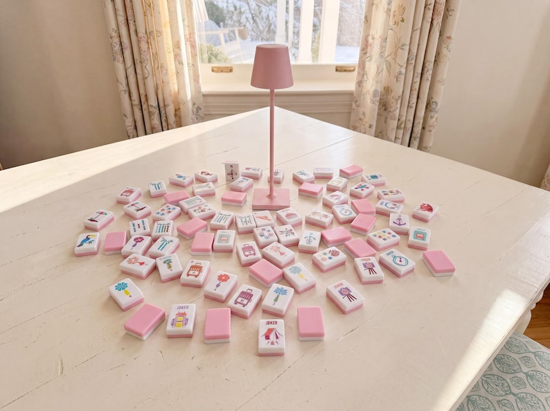 May include: A set of rectangular mahjong tiles with white faces featuring various colorful illustrations and pink backs, arranged in a scattered pattern around a tall, slender, solid pink table lamp with a conical shade on a white surface.