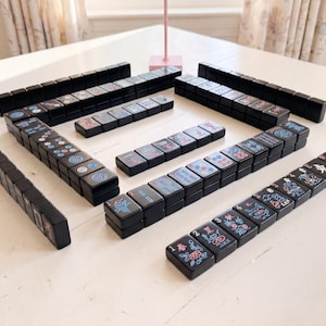 May include: A full Mahjong set with black tiles displaying colourful symbols and characters. The tiles are arranged on a white surface, ready for play. The game set includes multiple stacks of tiles, ideal for a game night.