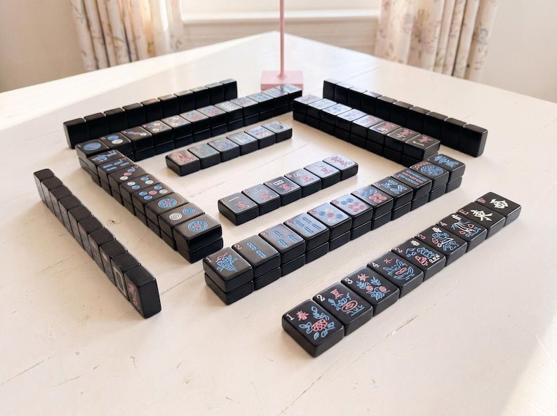 May include: A full Mahjong set with black tiles displaying colourful symbols and characters. The tiles are arranged on a white surface, ready for play. The game set includes multiple stacks of tiles, ideal for a game night.