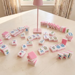 May include: A collection of pink and white Mahjong tiles with various symbols and characters scattered on a white table. A small pink lamp stands in the background. The tiles are arranged for play.