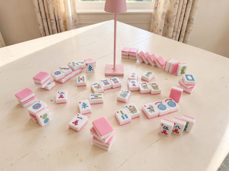 May include: A collection of pink and white Mahjong tiles with various symbols and characters scattered on a white table. A small pink lamp stands in the background. The tiles are arranged for play.