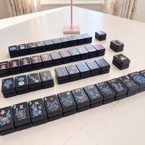 May include: A collection of black Mahjong tiles, displaying a variety of symbols and characters in red, blue, and white. The tiles are arranged on a white surface, demonstrating the game's layout. The set includes tiles with numbers, suits, and special characters.